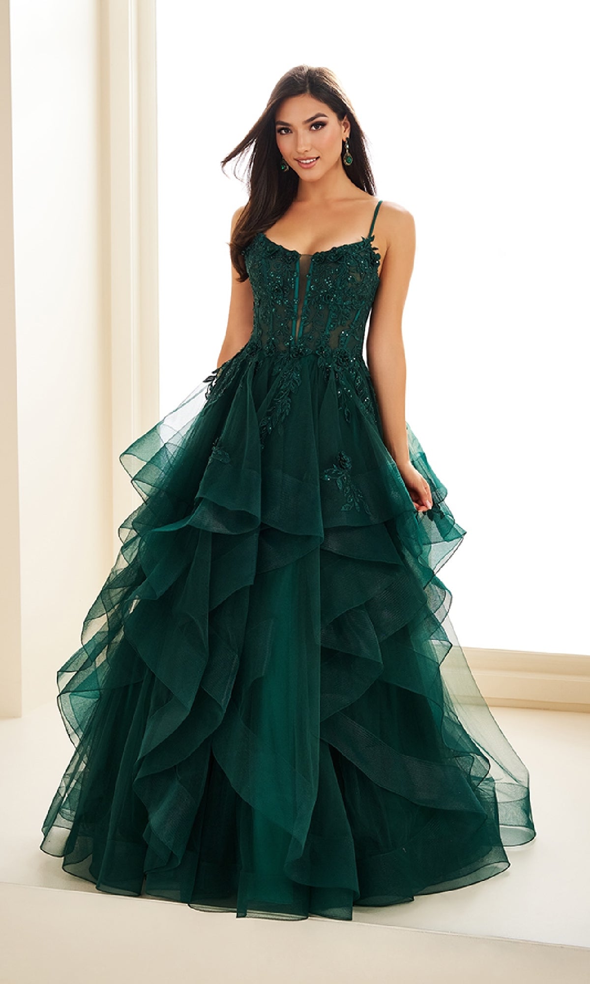 A woman stands indoors against a light background, wearing the Ellie Wilde EW36225 long prom dress—a dark green, tiered tulle ball gown with floral embellishments and thin straps.